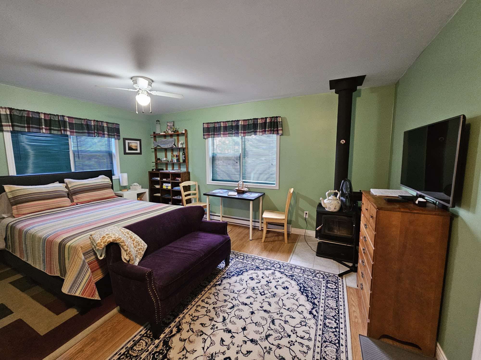 Studio room with queen bed, purple loveseat, dining table, wood stove and television